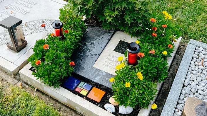 Das Grab von Sebastian und Max Podewilt. Auf dem Grab sind eine grüne Pflanze, Blumen, bunte Fliesen und zwei Grablichter.