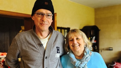 Stefan Becker, ein Mann mit silberner Brille, grauem Pullover und schwarzer Mütze, und Monika Keila, eine blondhaarigen Frau im blauen Pullover, stehen nebeneinander und lächeln freundlich.