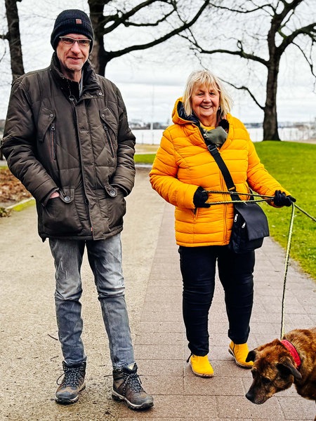 Stefan Becker und Monika Keiler, ein Mann und eine Frau mittleren Alters, spazieren mit zwei Hunden im Park.