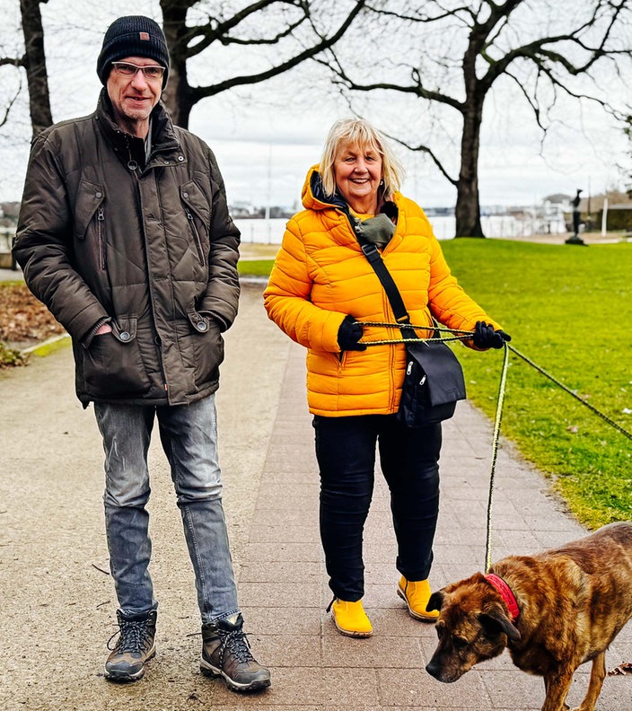 Stefan Becker und Monika Keiler, ein Mann und eine Frau mittleren Alters, spazieren mit zwei Hunden im Park.