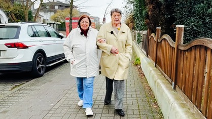 Zwei Frauen gehen einen Weg entlang, sie haben einander untergehakt