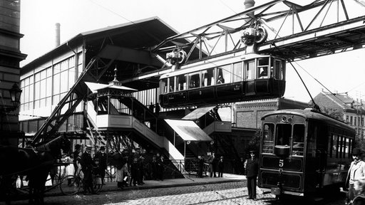 Schwarz-weiß Foto der Wuppertaler Schwebebahn aus dem Jahr 1901, die Bahn fährt aus dem Bahnhof Breitestraße heraus, unten auf der Straße stehen Menschen und es fährt eine elektrische Straßenbahn