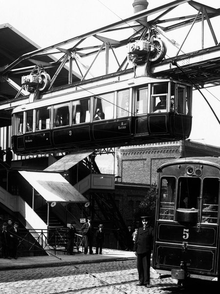 Schwarz-weiß Foto der Wuppertaler Schwebebahn aus dem Jahr 1901, die Bahn fährt aus dem Bahnhof Breitestraße heraus, unten auf der Straße stehen Menschen und es fährt eine elektrische Straßenbahn