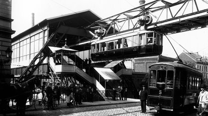 Schwarz-weiß Foto der Wuppertaler Schwebebahn aus dem Jahr 1901, die Bahn fährt aus dem Bahnhof Breitestraße heraus, unten auf der Straße stehen Menschen und es fährt eine elektrische Straßenbahn