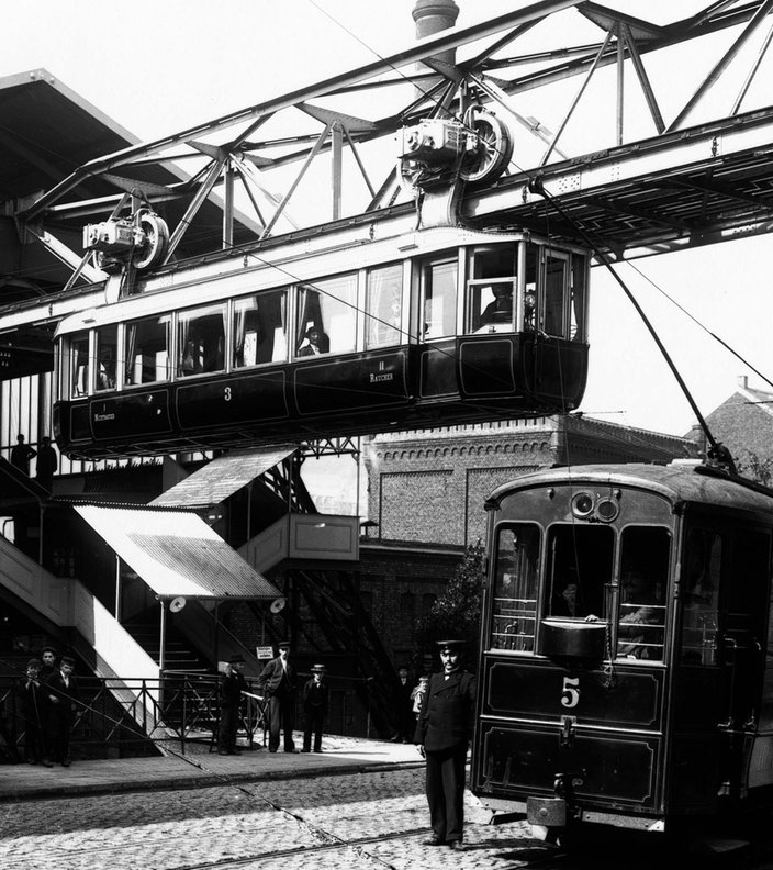 Schwarz-weiß Foto der Wuppertaler Schwebebahn aus dem Jahr 1901, die Bahn fährt aus dem Bahnhof Breitestraße heraus, unten auf der Straße stehen Menschen und es fährt eine elektrische Straßenbahn