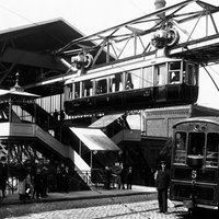 Schwarz-weiß Foto der Wuppertaler Schwebebahn aus dem Jahr 1901, die Bahn fährt aus dem Bahnhof Breitestraße heraus, unten auf der Straße stehen Menschen und es fährt eine elektrische Straßenbahn