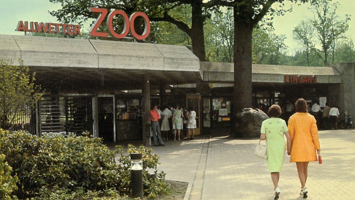 Alte Aufnahme aus dem Allwetterzoo Münster. Zwei Frauen mit einem mintgrünen und orangenen Kostüm gehen Richtung Eingang des Zoos. 