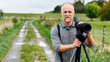 Ralph Achenbach steht auf einem Feldweg und stütz sich mit beiden Händen auf sein Spektiv, das auf einem Stativ steht. Er trägt ein gemustertes Hemd und einen Rucksack.