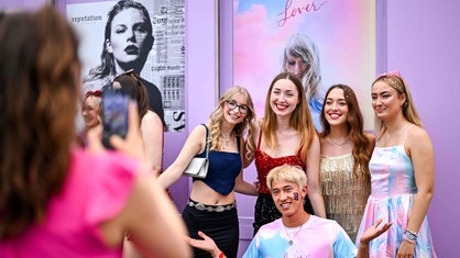 Fans von Taylor Swift machen Fotos in bunten Kostümen vor einem Plakat mit Taylor.