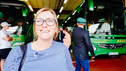 Ana Radermacher steht vor zwei Reisebussen, neben denen mehrere Menschen stehen. Sie lächelt in Richtung Kamera.