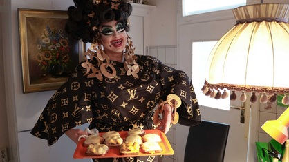 Andreas Patermann steht in seinem Freifrau von Kö Outfit in seiner Küche. Er trägt riesige goldene Ohringe und ein schwarzes Kleid. In beiden Händen hält er lächelnd ein Blech mit Toast Hawaii.