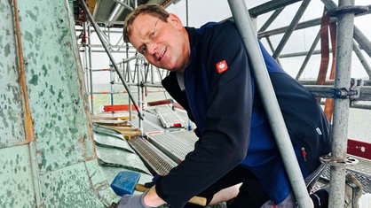 Gerald Plenter hält Werkzeug in beiden Händen, um die neuen Dachziegel zu bearbeiten. Er befindet sich oben auf dem Turm des Raesfelder Schlosses.