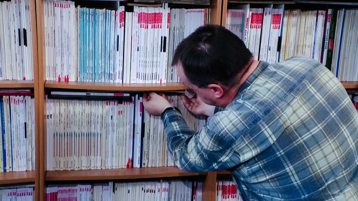 Andreas Niemann von hinten in kariertem Hemd vor einem Bücherregal, in das er gerade ein Heft reinlegt.