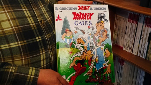 Zwei Hände halten ein Asterix-Comic in die Höhe, die Person trägt ein kariertes Hemd, rechts im Hintergrund sieht man ein Bücherregal.