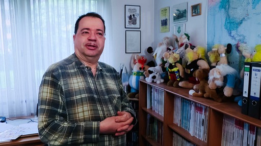 Andreas Niemann mit Brille und schütterem Haar in kariertem Hemd, die Hände vor seinem Bauch gefaltet, rechts hinter ihm Plüsch-Figuren aus den Asterix-Comics auf einem Bücherregal.