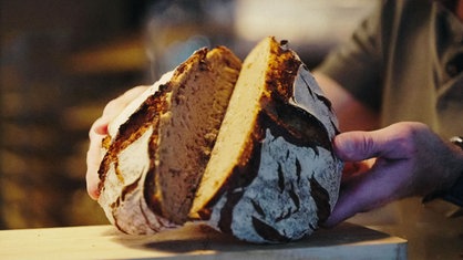 Zwei Hände halten ein frischgebackenes, dampfendes Brot, das in der Mitte aufgeschnitten ist.
