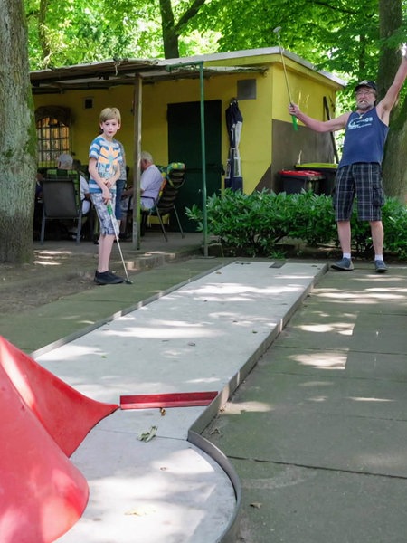 Ein Mann und ein Kind stehen gemeinsam vor einer Minigolfbahn. Beide haben Minigolfschläger in der Hand. 