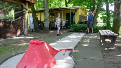 Ein Mann und ein Kind stehen gemeinsam vor einer Minigolfbahn. Beide haben Minigolfschläger in der Hand. 