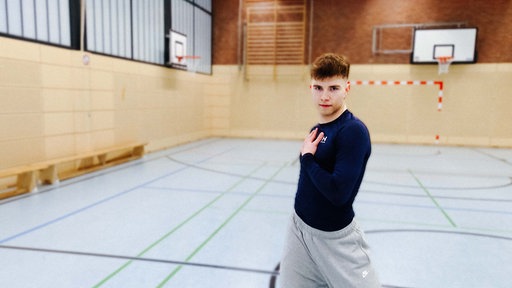 Tänzer Christian Weiß steht mit einer Hand auf der Brust in einer Turnhalle und schaut in die Kamera.