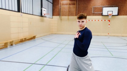 Tänzer Christian Weiß steht mit einer Hand auf der Brust in einer Turnhalle und schaut in die Kamera.