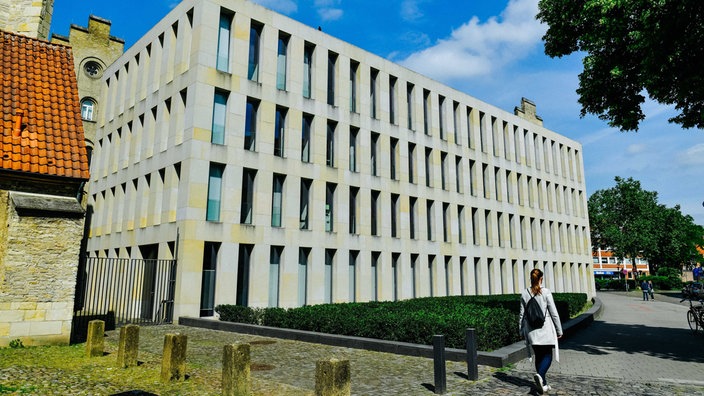 Frau mit schwarzem Tunrbeutel läuft an Diözesanbibliothek in Münster vorbei, die aus beigem Sandstein und rechteckigen, senkrechten Öffnungen besteht
