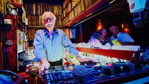 DJ Dietmar steht hinter seinem DJ-Pult und lächelt in die Kamera. Im Hintergrund stehen zahlreiche Schallplatten.