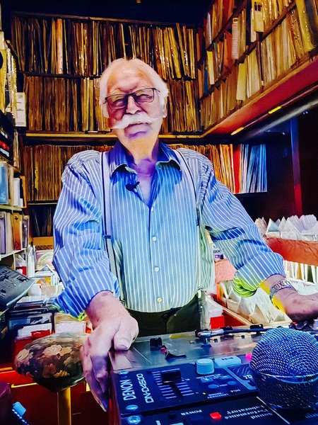 DJ Dietmar steht hinter seinem DJ-Pult und lächelt in die Kamera. Im Hintergrund stehen zahlreiche Schallplatten.