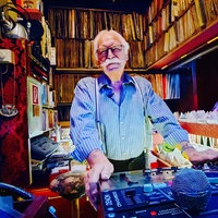 DJ Dietmar steht hinter seinem DJ-Pult und lächelt in die Kamera. Im Hintergrund stehen zahlreiche Schallplatten.