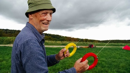 Christoph Dünnwald, blaues Karohemd, kaki-farbene Mütze, lächelt in die Kamera, in seinen Händen hält er einen roten und einen gelben Ring, an dem weiße Fäden in die Luft gehen.