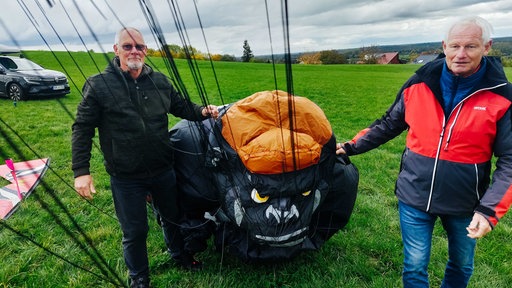 Jürgen Jansen (rechts) und Hans Schneider (links) stehen auf einer grünen Wiese und halten einen orange-schwarzen Stoff mit dem Gesicht eines Gorillas.