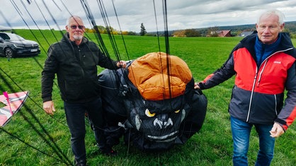 Jürgen Jansen (rechts) und Hans Schneider (links) stehen auf einer grünen Wiese und halten einen orange-schwarzen Stoff mit dem Gesicht eines Gorillas.