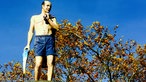 Ein Säulenheiliger in Badehose steht auf einer Litfaßsäule.
