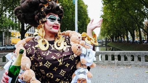 Die Freifrau von Kö posiert auf einer Brücke der Düsseldorfer Königsallee. Sie trägt ein schwarzes Kleid und große goldene Ohrrigne und Ketten. In der rechten Hand hält sie eine offenene Sektflasche. 