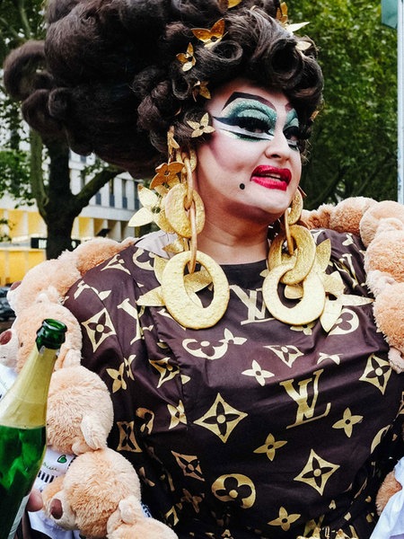 Die Freifrau von Kö posiert auf einer Brücke der Düsseldorfer Königsallee. Sie trägt ein schwarzes Kleid und große goldene Ohrrigne und Ketten. In der rechten Hand hält sie eine offenene Sektflasche. 