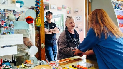 Verena Kirchberg steht in ihrem Büdchen und greift über die Verkaufstheke die Hand einer Kundin. Im Hintergrund lächelt ein weiterer Kunde.