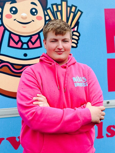 Noel Schnieders steht in einem pinken Pullover vor seinem hellblauen Imbisswagen