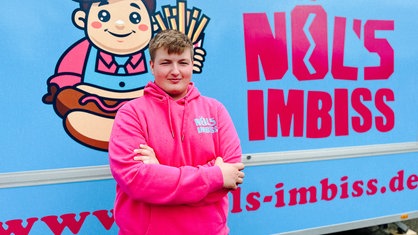 Noel Schnieders steht in einem pinken Pullover vor seinem hellblauen Imbisswagen