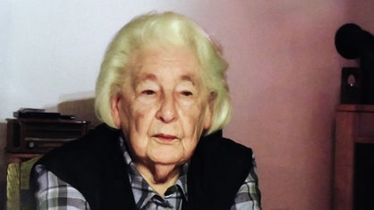 Eine ältere Frau mit grauen Haaren und karierter Bluse schaut ernst