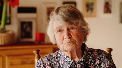 Eine ältere Frau mit grauen Haaren und Blumenbluse schaut ernst in Richtung Kamera