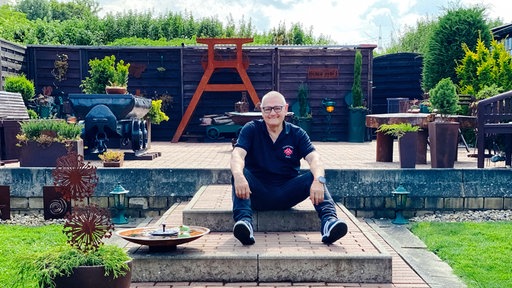 Ein älterer Mann sitzt auf einer Stufe in seinem Garten. Im Hintergrund stehen eine Lore und ein Förderturm.