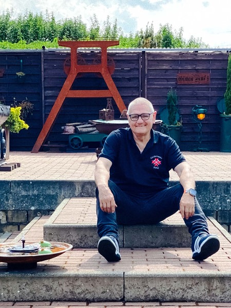 Ein älterer Mann sitzt auf einer Stufe in seinem Garten. Im Hintergrund stehen eine Lore und ein Förderturm.