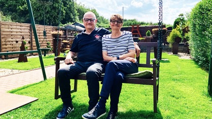 Ein älterer Mann und eine ältere Frau sitzen auf einer Bank in ihrem Garten und lächeln in die Kamera.