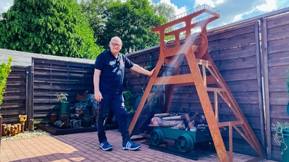 Ein älterer Mann steht neben einem rotbraunen Förderturm, der etwas größer ist als er selbst.