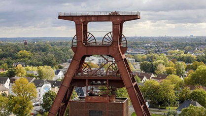 Der Doppelblock der Zeche Zollverein in Essen, dahinter ein weiter Blick über Häuser und Bäume in der Stadt.