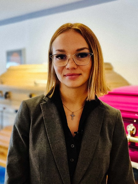 Eine junge Bestatterin mit kurzen Haaren und Brille schaut neutral in die Kamera. Im Hintergrund sind einige Särge.