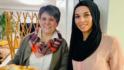 Eine Frau mit gräulichen kurzen Haaren und Brille steht neben einer Frau mit Kopftuch und rosa Bluse.
