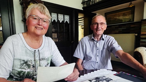 Ingrid und Arnold Arendsen, sie kurzes blondes Haar und Brille mit weiß-grau-kariertem Hemd, hält ein Blatt in der Hand, er kurze graue Haare, Brille, blau-weiß-kariertes Hemd, sitzen am Tisch.