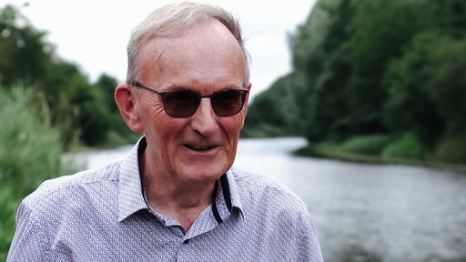 Arnold Arendsen, kurze graue Haare, braun getönte Sonnenbrille, weiß-blau-kariertes Hemd, hinter ihm fließt ein Fluss.