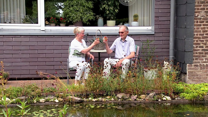Ehepaar Arendsen sitzt auf zwei Stühlen an einem Tisch in ihrem Garten. Im Vordergrund ein Teich.