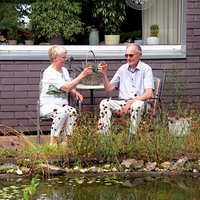 Ehepaar Arendsen sitzt auf zwei Stühlen an einem Tisch in ihrem Garten. Im Vordergrund ein Teich.